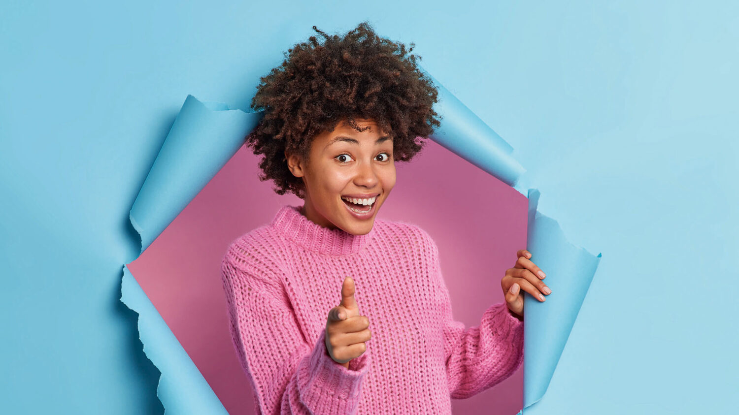 Positive beautiful curly ethnic woman makes you got this gesture praises good job points at you encourages person smiles gladfully wears knitted sweater breaks through blue paper background.