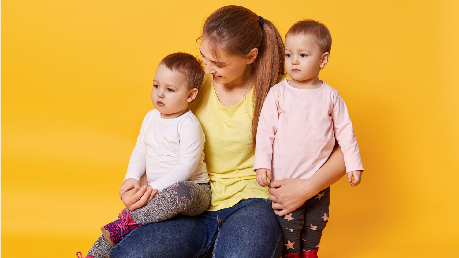 A smiling woman sits with her fair-haired daughters, hugs kids and feels happy. Lovely children look away attentively, spend time with their mother, play together. Family, children and care concept.